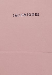 Lichtroze katoenen kledingstuk met de tekst "JACK&JONES" geborduurd in donkerblauw, weergegeven op een gladde, textuurachtige stofoppervlak.