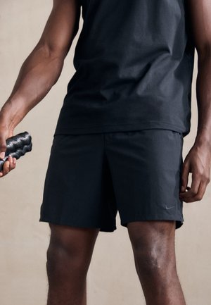 Homme portant un short et un t-shirt de sport noirs, tenant un rouleau de massage texturé noir dans sa main gauche.