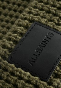 Oliveno zelená tkanina s textúrou, na ktorej je čierny kožený štítok s embosovaným nápisom "ALLSAINTS". Materiál vyzerá hrubo a tkane.