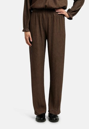 Kobieta w brązowych, teksturowanych długich spodniach i dopasowanym topie, stojąca z jedną ręką na biodrze, w czarnych butach na białym tle.