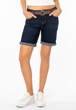 Jeans-Shorts, dunkelblau, mit umgeschlagenem Saum und kontrastierenden Nähten. Brauner Gürtel mit einer Metallschnalle. Fünf-Taschen-Design. Weiße Sneakers.