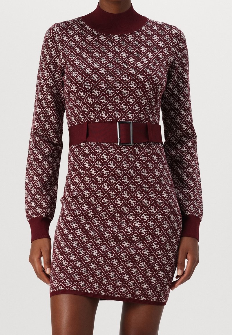 Femme portant une robe en maille bordeaux ajustée avec des motifs en losange blancs, manches longues, col montant et une ceinture assortie avec une boucle argentée.