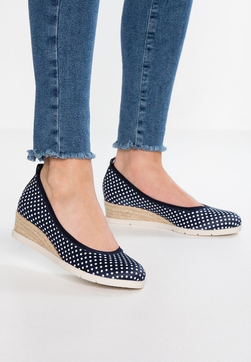 Pieds chaussés de chaussures compensées bleu marine à pois avec talons tissés beige et semelles blanches, associés à un jean skinny bleu à ourlet effiloché.