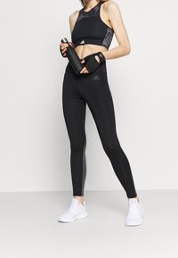 Musta urheilusali, jossa on kaarrokkeet, joissa on kuvioita, yhdistetty mustiin legginsseihin, joissa on hienovarainen sivutekstuuri. Vesipullo täydentää asun.