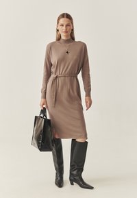 Neulemekko taupe-värissä, jossa vyötärösolki, pitkät hihat ja sivuhalkiot. Yhdistetty mustiin polven korkeisiin saappaisiin ja kiiltävään mustaan olkalaukkuun.