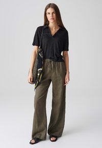 Polo a maniche corte nera abbinata a pantaloni larghi in lino verde oliva. Il modello tiene una sciarpa fantasia e una borsa intrecciata. Ai piedi sandali neri piatti.