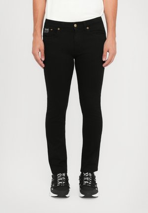 Black skinny jeans with a smooth texture, featuring two front pockets, a coin pocket, and gold button hardware. Worn with black sneakers.