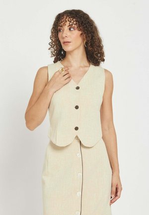 Mujer con cabello rizado, vestida con chaleco beige abotonado y falda, complementada con anillo y pendientes de oro, mirando de lado.