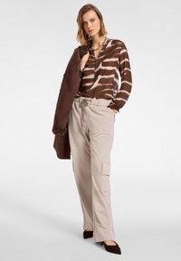 Braun gemustertes Hemd mit langen Ärmeln, beigefarbene Cargo-Hosen und dunkelbraune spitze Ballerinas. Das Hemd hat ein Batikdesign.