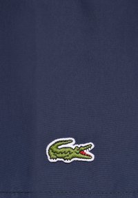 Marineblauer Stoff mit einer glatten Textur zeigt ein grünes Krokodil-Logo, das in Weiß umrandet ist und sich in der unteren rechten Ecke befindet.