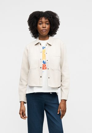 Junge Frau mit natürlichen lockigen Haaren, die eine hellbeige Jacke mit Knöpfen über einem weißen Grafik-T-Shirt und dunkelblauen Jeans trägt.