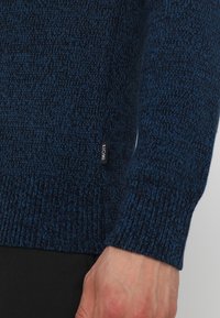 Nära bild av en marinblå stickad tröjas ärm och midja med en liten svart KIOMI-märke, bärs av en person med en synlig hand.