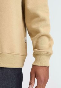 Beige sweatshirt made of soft fabric. Features ribbed cuffs and hem, with a small brand tag on the side. It's relaxed fit with a smooth texture.