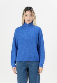 Pulover tricotat albastru cu guler înalt și închidere cu fermoar pe jumătate, având un model texturat cu nervuri și mâneci lungi. Purtați cu blugi din denim.
