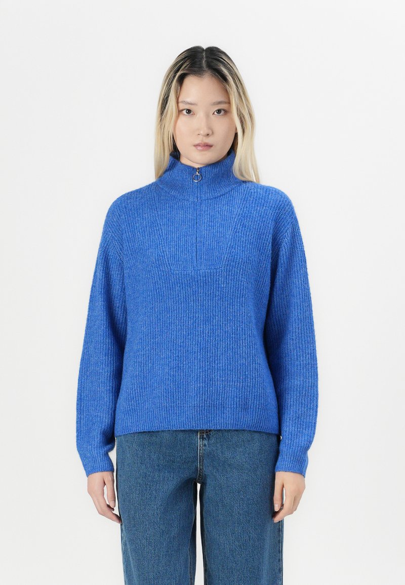 Pulover tricotat albastru cu guler înalt și închidere cu fermoar pe jumătate, având un model texturat cu nervuri și mâneci lungi. Purtați cu blugi din denim.