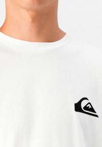 Persona che indossa una maglietta bianca con un piccolo logo Quiksilver nero sul petto sinistro, mostrata dal collo a metà torso.