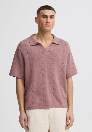 Mann mit kurzen dunklen Haaren, der ein mauvefarbenes, gestricktes Poloshirt mit kurzem Ärmel und Rautenmuster sowie beige Hosen trägt.