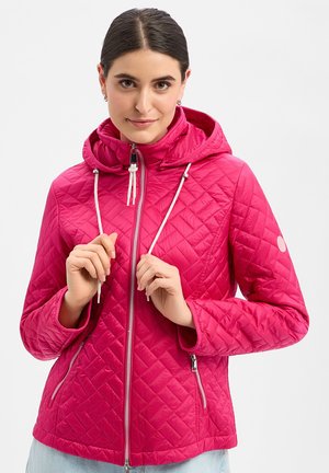 Vrouw past de trekkoorden aan van een felroze gewatteerde jas met capuchon en rits, draagt lichtblauwe spijkerbroek, tegen een effen achtergrond.