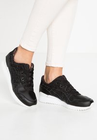 Svarta sneakers med en glänsande lädertåvidd, texturerat svart tygöverkinn och vita gummisulor. Snörningsdesign med subtila färgglada detaljer.