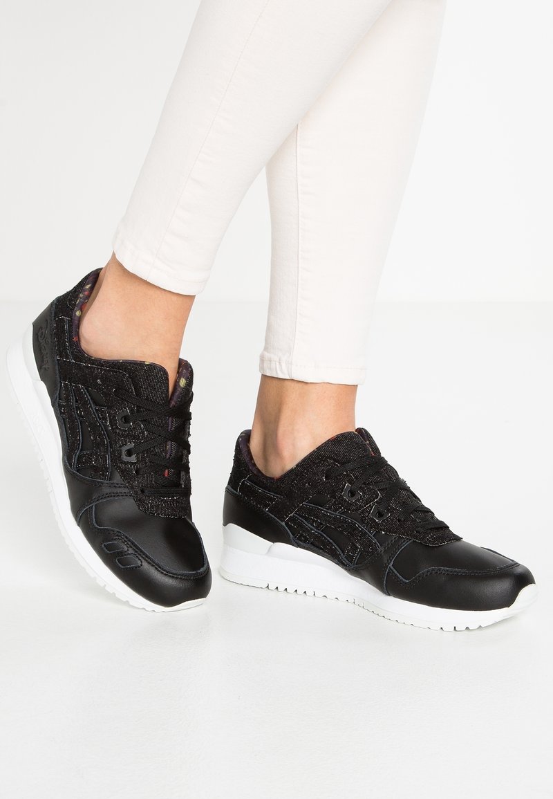 Svarta sneakers med en glänsande lädertåvidd, texturerat svart tygöverkinn och vita gummisulor. Snörningsdesign med subtila färgglada detaljer.