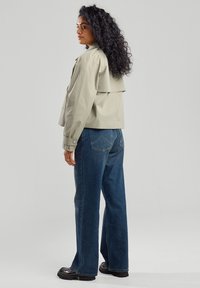 Vaaleanharmaa lyhyt takki, jossa on kerroksellinen selkäosa, yhdistettynä leveälahkeisiin tummansinisiin farkkuihin ja mustiin korkokenkiin. Teksturoitu hiustyyli näkyvissä.