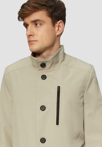 Lichtbeige jas van zacht materiaal, met een hoge kraag, knoopsluiting aan de voorkant en een ritszak voor extra opbergruimte.