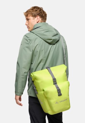Man draagt een groene capuchonjas en heeft een fluorescerend gele waterdichte Vaude roll-top rugzak op zijn rug.