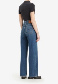 Femme portant un jean bleu taille haute à jambes larges et un top noir à manches courtes, debout de profil avec une main détendue le long du corps.
