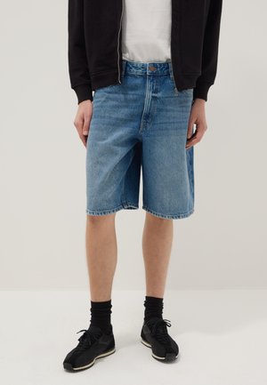 Persona che indossa pantaloncini di jeans azzurri di media lunghezza, scarpe da ginnastica nere con calzini neri, maglietta bianca e giacca nera con cerniera.