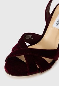 Sandal s visokom petom od burgundske baršunaste tkanine s prekriženim trakama, glatkim prednjim dijelom i laganim unutrašnjim podstavom. Zatvorena stražnja strana za sigurno prianjanje.