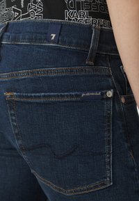 Los jeans de mezclilla azul oscuro presentan una cinturilla alta, costuras amarillas en contraste y una etiqueta que dice "for all mankind" cerca del bolsillo trasero.