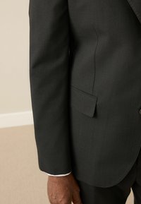 Black tailored suit jacket featuring a smooth texture, single breast pocket, and visible white cuff. Close-up view of the sleeve.