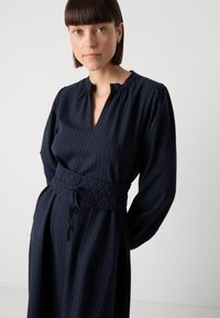 Robe bleu marine à rayures verticales, avec un col plissé et une taille élastique texturée dotée d'un détail de cordon de serrage. Manches longues.