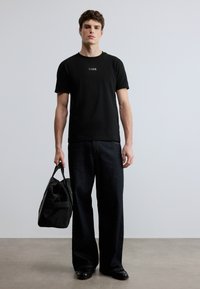 Μαύρο βαμβακερό μπλουζάκι με λογότυπο, συνδυασμένο με φαρδιά σκούρα τζιν. Το μοντέλο κρατά μια μαύρη υφασμάτινη τσάντα, στέκεται σε ανοιχτό γκρι δάπεδο.