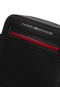 Μαύρο τσάντα χιαστί με υφή, με το λογότυπο Tommy Hilfiger και χαρακτηριστικές κόκκινες, λευκές και μπλε ρίγες στο μπροστινό μέρος.