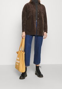 Chaqueta de camisa de pana marrón con dos bolsillos, llevada sobre un suéter de cuello alto oscuro, combinada con jeans azules recortados y botas negras con cordones. Sosteniendo una bolsa de mano amarilla.