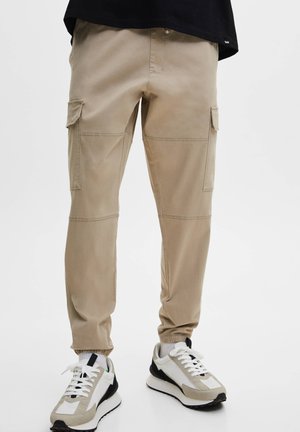Beige Cargo-Hosen mit Knie-Nähten und elastischen Bündchen, getragen mit weißen, beige-farbenen und schwarzen Sneakers, einer Person, die vor einem weißen Hintergrund steht.