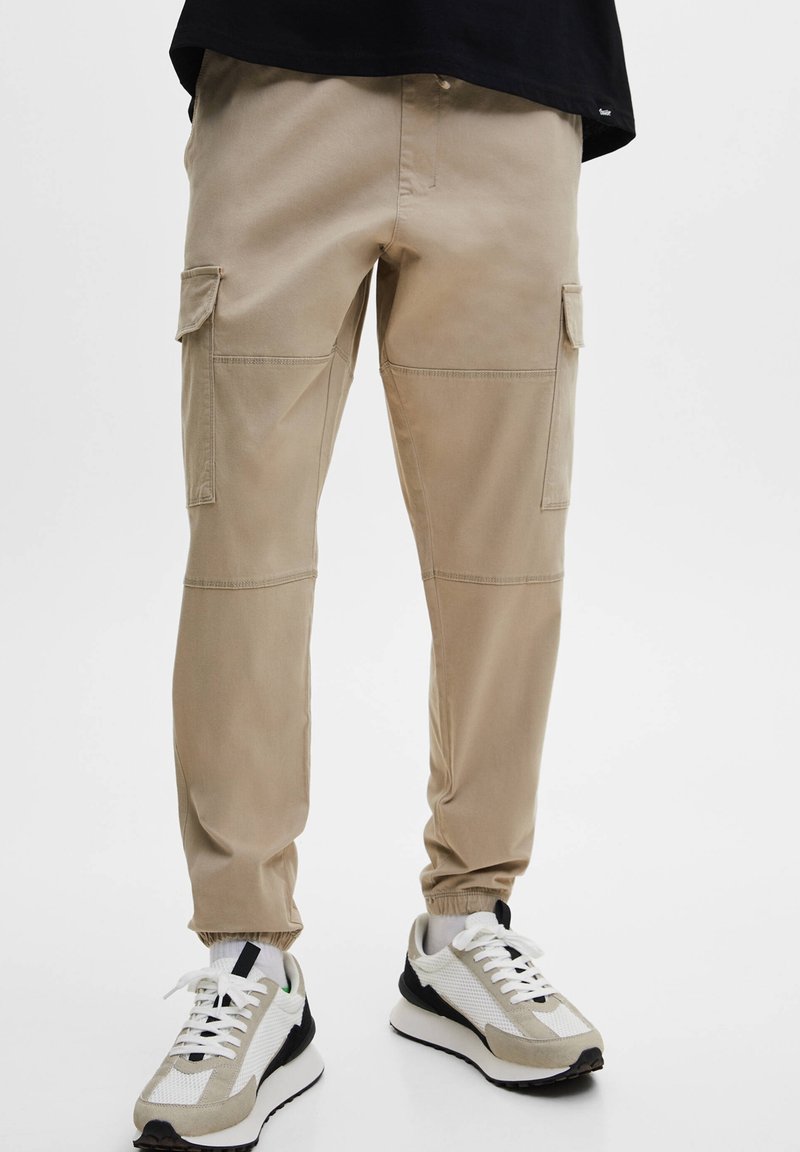 Beige Cargo-Hosen mit Knie-Nähten und elastischen Bündchen, getragen mit weißen, beige-farbenen und schwarzen Sneakers, einer Person, die vor einem weißen Hintergrund steht.