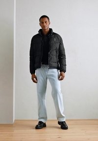 Svart polstret jakke med høy krage, svart polo-skjorte, lyseblå jeans og svarte lærskor, stående mot en nøytral vegg.