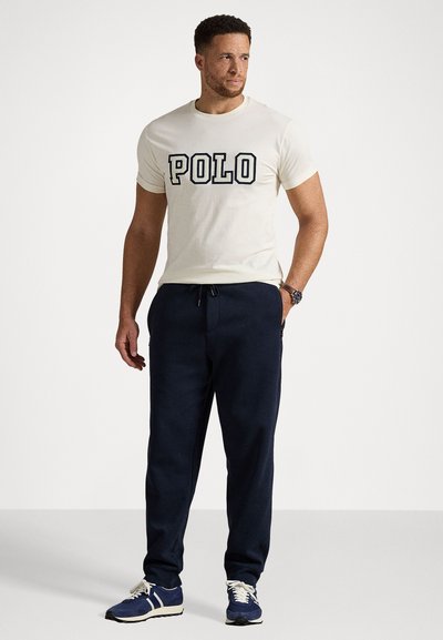 Krémové tričko s grafikou "POLO", spárované s námořnickými tepláky. Boty jsou modré s bílými akcenty. Neformální sportovní outfit.