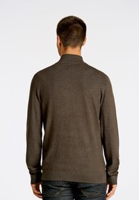 Maglione a maglia marrone scuro con colletto alto e maniche lunghe, caratterizzato da un orlo e polsini a coste, indossato con jeans in denim scuro.