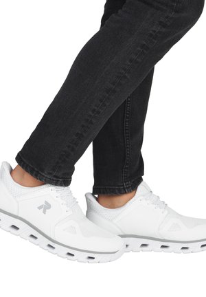 Personne portant des baskets blanches de sport avec des panneaux en mesh et un jean noir, jambes croisées aux chevilles sur un fond blanc.