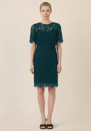 Frau mit blonden Haaren in einem knieumspielenden, dunkelgrünen Spitzenkleid und schwarzen offenen Schuhen, stehend vor einem beigen Hintergrund, nach vorne blickend.