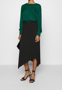 Blusa verde a maniche lunghe in tessuto morbido; gonna midi nera plissettata con orlo asimmetrico; tacchi neri con cinturini e punta quadrata.