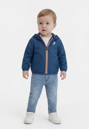 Malé dieťa oblečené v modrej kapucovej bunde, svetlomodrých džínsoch a bielych teniskách, stojace na bielom pozadí.