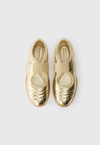 Zelta metāliskas baleta kurpes ar kvadrātveida struktūru, elastīgu siksniņu pāri pēdas vidum un mīkstinātu iekšējo zoli, kas aprīkota ar brūnu gumijas ārējo zoli.