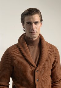 Cárdigan de canalé marrón con cuello de chal, que presenta un tejido texturizado y dos botones. Lleva puesto sobre un cuello alto a juego.