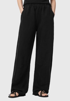 Pantalon noir ample avec taille élastique et poches latérales, porté avec des sandales noires sur un fond clair. Mains dans les poches.