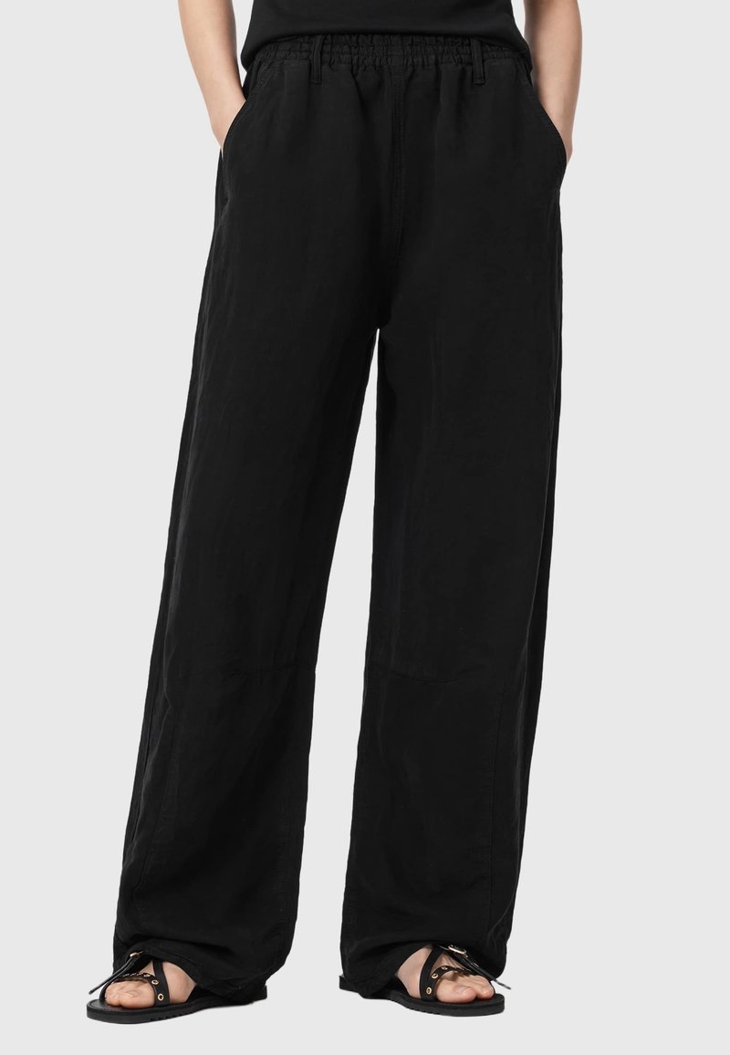 Black loose-fit pants with elastic waistband and side pockets, worn with black sandals on a light background. Hands in pockets.
