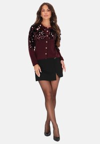 Donna che indossa un cardigan bordeaux decorato con paillettes, una mini gonna nera con spacco frontale, collant trasparenti e tacchi neri, in piedi con le gambe incrociate.